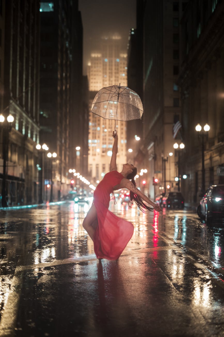 woman holding umbrella dancing in the middle of the road near cars and buildings